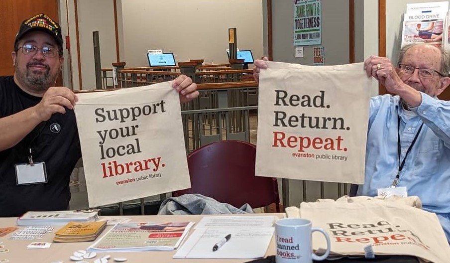 Two people sitting at a table, holding up library bags that say "Support Your Local Library" and "Read, Return, Report."